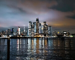 Nighttime panorama of Lower Manhattan's illuminated skyline, captured from Weehawken's riverfront with foreground pilings visible