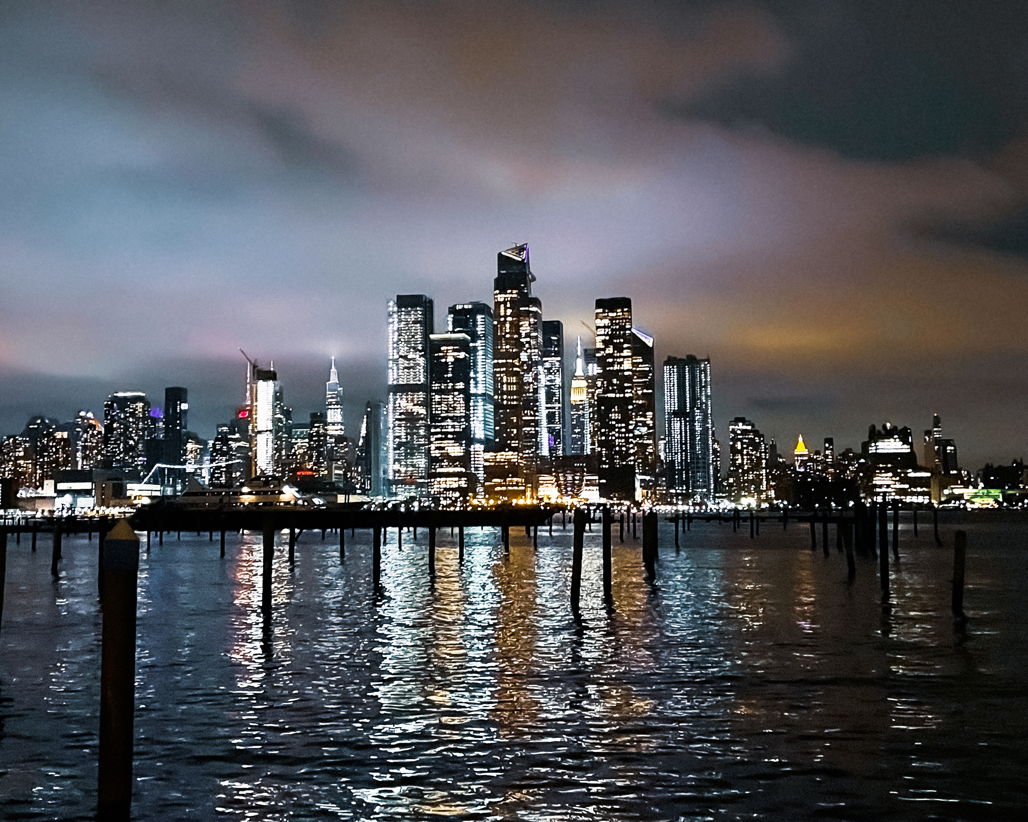 Nighttime panorama of Lower Manhattan's illuminated skyline, captured from Weehawken's riverfront with foreground pilings visible
