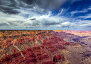 Panoramic view of the Grand Canyon’s vivid red and orange cliffs, sharply textured in bright light, with a turbulent sky of dark storm clouds and lighter breaks above.
