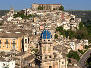 The blue dome of Santa Maria dell’Itria emerging above the layered rooftops of Ragusa Ibla, captured in soft Mediterranean light.