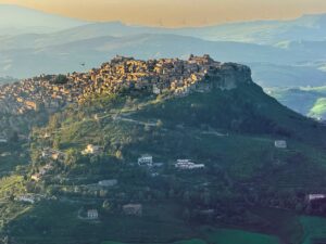 Sunlit stone buildings of Calascibetta atop a steep cliff in central Sicily during sunset, viewed from Enna with misty distant peaks.